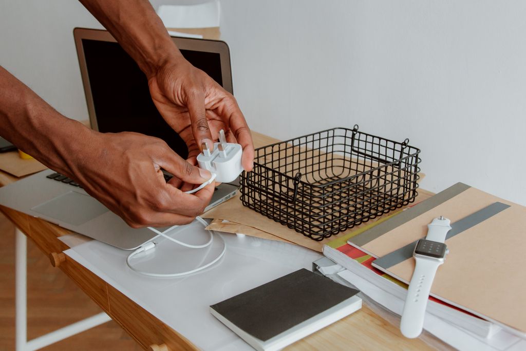 Close-up of a man organizing a desk, handling a charger next to a laptop, wire basket, and notebooks.