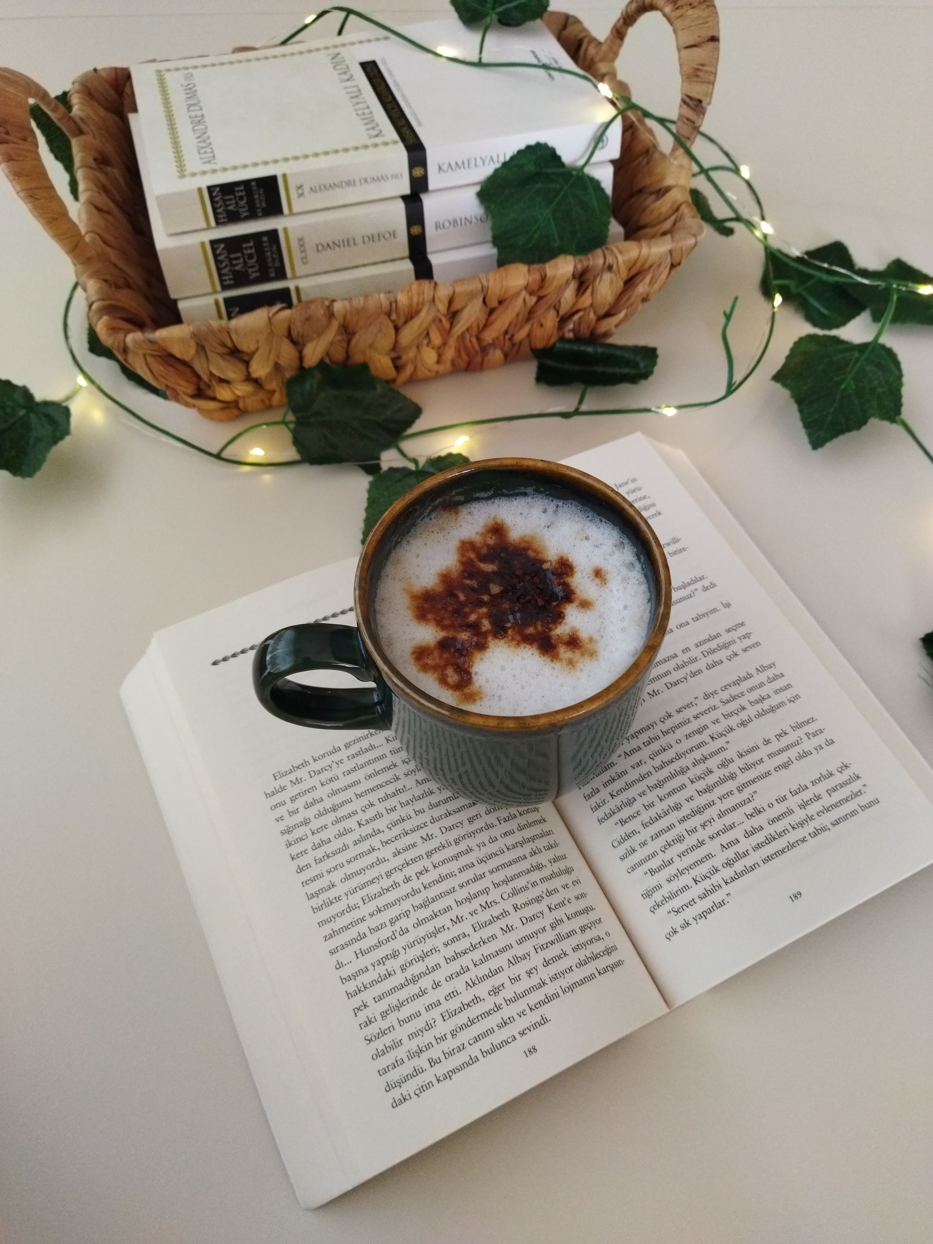 A warm cappuccino on an open book, surrounded by a basket of books and greenery.